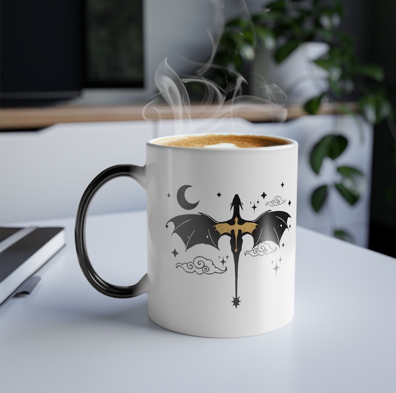 May include: A white ceramic mug with a black handle. The mug features a black and gold dragon silhouette flying against a white background with clouds and stars. The mug is filled with coffee.