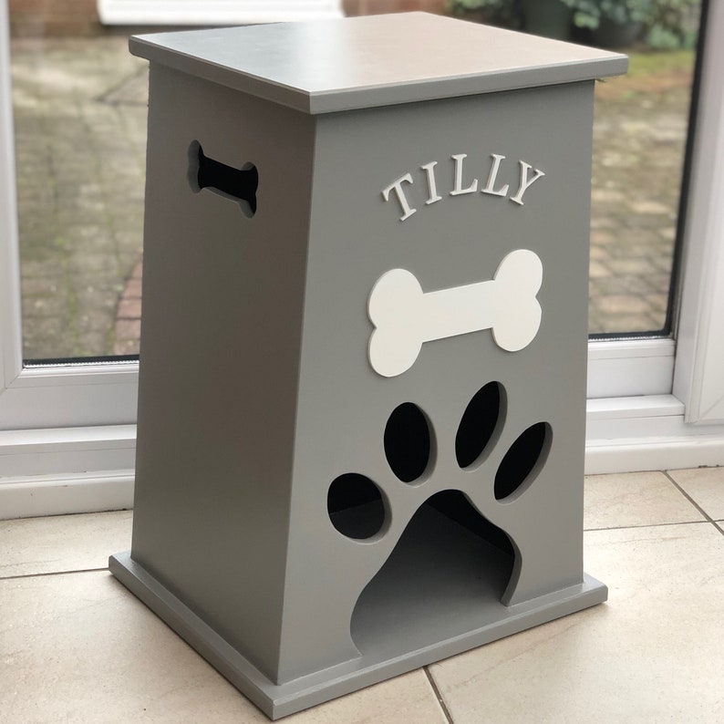 Peut inclure: Un conteneur de stockage de nourriture pour animaux de compagnie en bois gris avec une d&eacute;coupe en forme d'os. Le conteneur pr&eacute;sente un os blanc et le nom "TILLY" au-dessus d'une d&eacute;coupe en forme de patte. Le conteneur est rectangulaire.