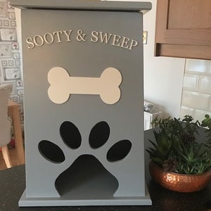Peut inclure: Un conteneur de stockage de nourriture pour animaux de compagnie en bois gris avec les mots "SOOTY & SWEEP" en lettres blanches. Il pr&eacute;sente un design en forme d'os et une d&eacute;coupe en forme de patte. Id&eacute;al pour les animaux.
