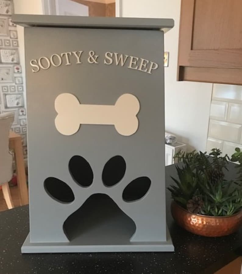 Peut inclure: Un conteneur de stockage de nourriture pour animaux de compagnie en bois gris avec les mots "SOOTY & SWEEP" en lettres blanches. Il pr&eacute;sente un design en forme d'os et une d&eacute;coupe en forme de patte. Id&eacute;al pour les animaux.
