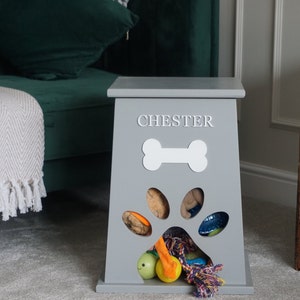 May include: Gray wooden dog toy storage bin with a paw print cutout and a bone shape on the front. The bin is labeled "CHESTER" and has a variety of dog toys inside.