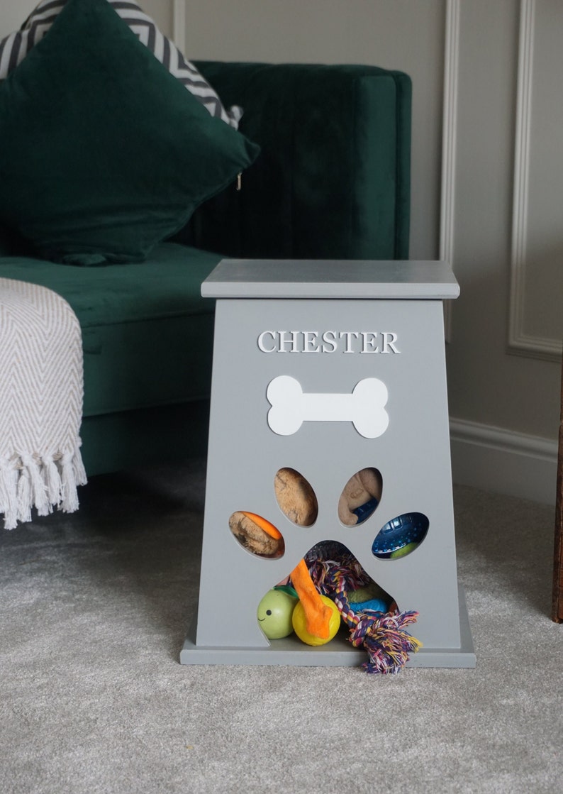 May include: Gray wooden dog toy storage bin with a paw print cutout and a bone shape on the front. The bin is labeled "CHESTER" and has a variety of dog toys inside.