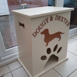 Peut inclure: Un conteneur de stockage de nourriture pour animaux de compagnie en bois cr&egrave;me avec les noms "DOUGY & DEXTER" en lettres marron. Il pr&eacute;sente une silhouette de teckel marron et une d&eacute;coupe en forme de patte. Il y a une d&eacute;coupe en forme d'os sur le c&ocirc;t&eacute;.