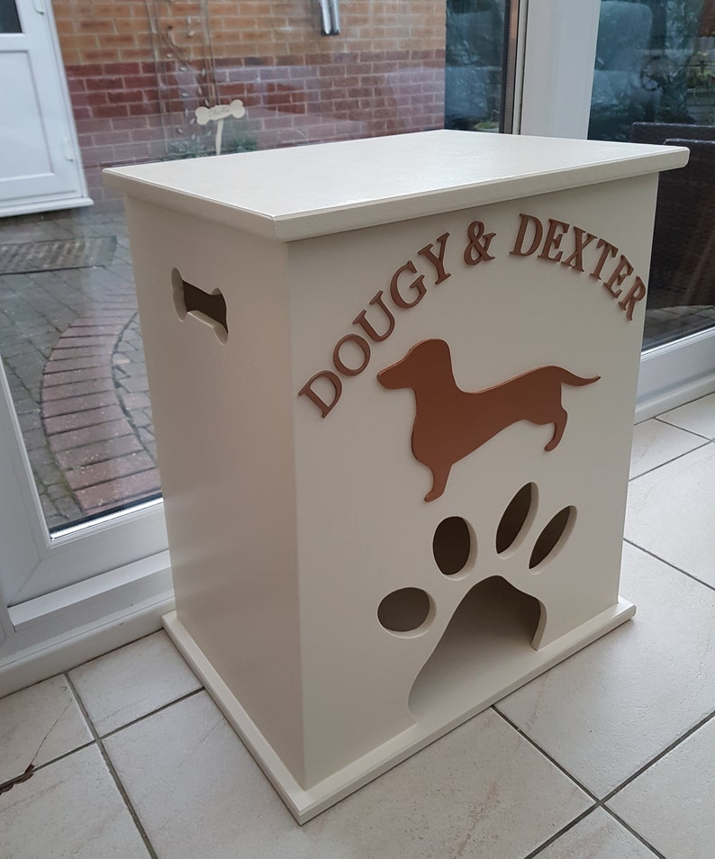 Peut inclure: Un conteneur de stockage de nourriture pour animaux de compagnie en bois cr&egrave;me avec les noms "DOUGY & DEXTER" en lettres marron. Il pr&eacute;sente une silhouette de teckel marron et une d&eacute;coupe en forme de patte. Il y a une d&eacute;coupe en forme d'os sur le c&ocirc;t&eacute;.