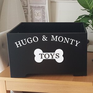 May include: Black wooden toy box with the names "HUGO & MONTY" in white script above a white bone-shaped graphic with the word "TOYS". The box is rectangular and sits on a wooden surface.