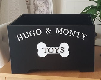 Peut inclure: Coffre à jouets en bois noir avec les noms "HUGO & MONTY" en écriture blanche au-dessus d'un os blanc avec le mot "TOYS". La boîte est rectangulaire et repose sur une surface en bois.