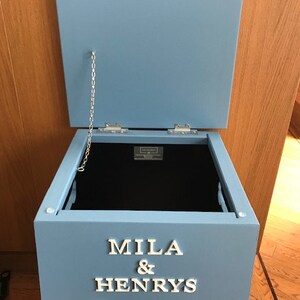Peut inclure: Un coffre &agrave; jouets en bois bleu clair avec un couvercle ouvert. Le coffre porte l'inscription blanche "MILA & HENRYS TOYS" et un motif en forme d'os. Une cha&icirc;ne argent&eacute;e relie le couvercle au coffre.