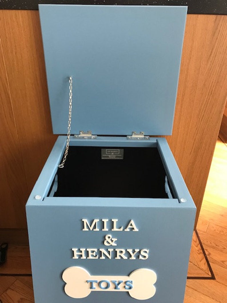 Peut inclure: Un coffre &agrave; jouets en bois bleu clair avec un couvercle ouvert. Le coffre porte l'inscription blanche "MILA & HENRYS TOYS" et un motif en forme d'os. Une cha&icirc;ne argent&eacute;e relie le couvercle au coffre.