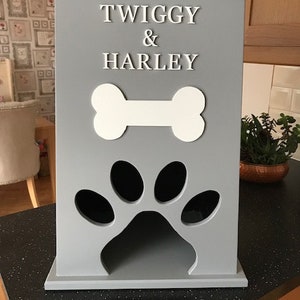 Peut inclure: Un r&eacute;cipient &agrave; friandises pour animaux de compagnie en bois gris avec les noms "TWIGGY & HARLEY" en lettres blanches. Il pr&eacute;sente un motif d'os blanc et une d&eacute;coupe en forme de patte. Le r&eacute;cipient est sur un comptoir noir.