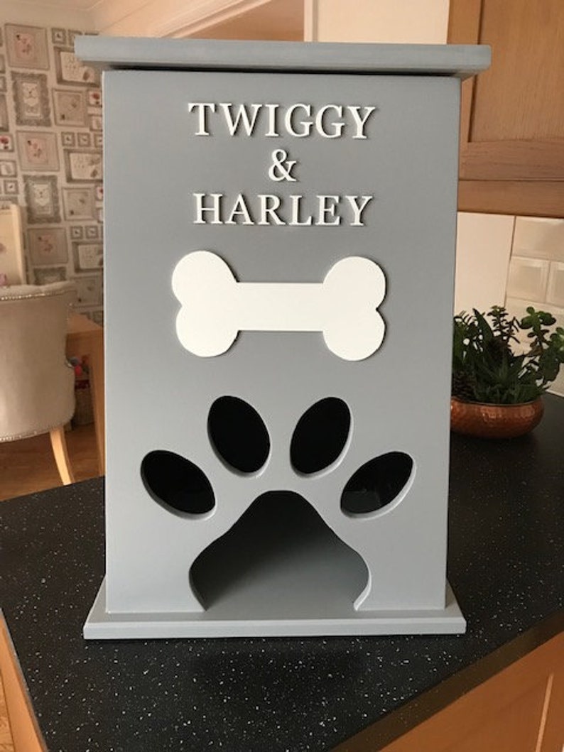 Peut inclure: Un r&eacute;cipient &agrave; friandises pour animaux de compagnie en bois gris avec les noms "TWIGGY & HARLEY" en lettres blanches. Il pr&eacute;sente un motif d'os blanc et une d&eacute;coupe en forme de patte. Le r&eacute;cipient est sur un comptoir noir.