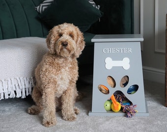Peut inclure: Un bac de rangement pour jouets de chien gris avec une découpe en forme de patte et le nom "CHESTER" sur le devant. Le bac est rempli de divers jouets pour chien, dont des cordes, des balles et des jouets en peluche. Un chien brun clair est assis devant le bac.