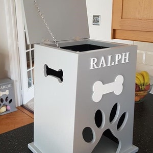 May include: A gray wooden dog treat storage bin with a white paw print and bone cutout. The bin has a hinged lid with a chain to keep it open. The bin is labeled "RALPH".