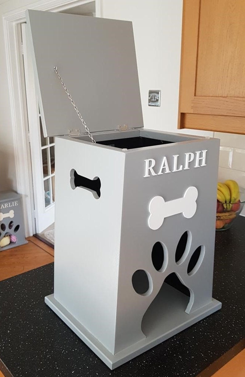 May include: A gray wooden dog treat storage bin with a white paw print and bone cutout. The bin has a hinged lid with a chain to keep it open. The bin is labeled "RALPH".