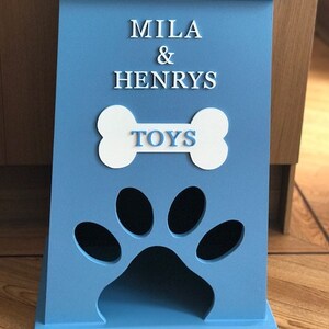 Peut inclure: Un coffre &agrave; jouets pour animaux de compagnie en bois bleu clair. Il pr&eacute;sente une d&eacute;coupe en forme de patte et les mots "MILA & HENRYS TOYS" en lettres blanches. Le coffre a un couvercle.