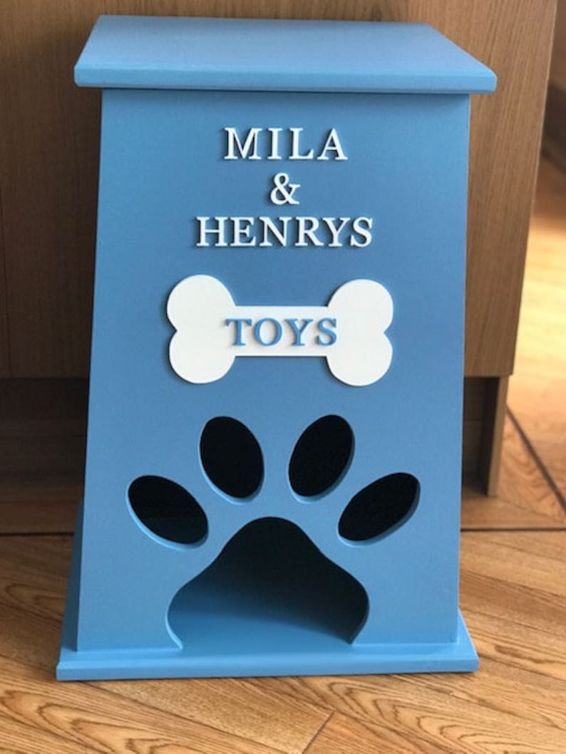 Peut inclure: Un coffre &agrave; jouets pour animaux de compagnie en bois bleu clair. Il pr&eacute;sente une d&eacute;coupe en forme de patte et les mots "MILA & HENRYS TOYS" en lettres blanches. Le coffre a un couvercle.