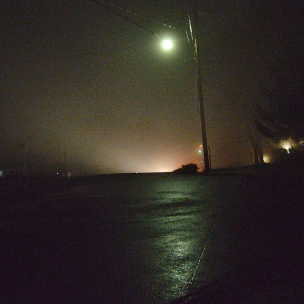 Eerie foggy night with lone street lamp and reflection on street and some house lights, mostly dark.