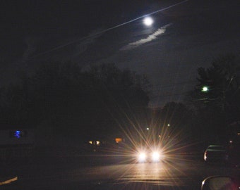 Night view of car with bright headlights driving down street with cloudy moonlit sky