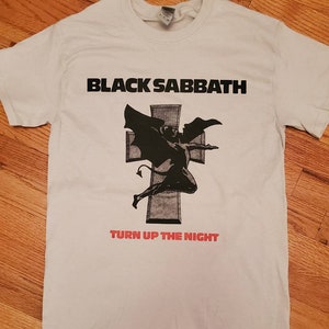 May include: White t-shirt with a black and white graphic of a winged demon on a cross. The text "BLACK SABBATH" is printed in black above the graphic and "TURN UP THE NIGHT" is printed in red below the graphic.