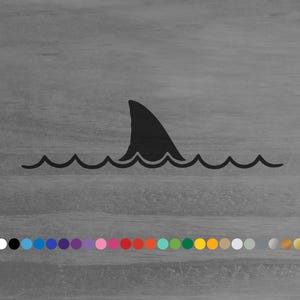 May include: A black graphic decal featuring a shark fin protruding above a stylized wavy line representing water. Below the main graphic, a horizontal row of twenty-two small circular color swatches is displayed in various shades and finishes.