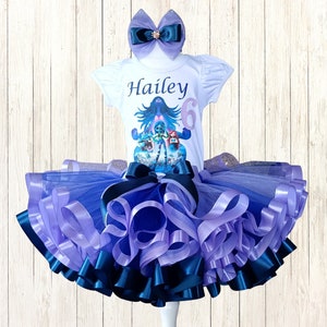 May include: A white short-sleeved shirt with the name "Hailey" and the number "6" printed on it, paired with a layered tutu skirt in shades of blue and purple. A matching bow with a jewel accent completes the set. The outfit is displayed on a mannequin.