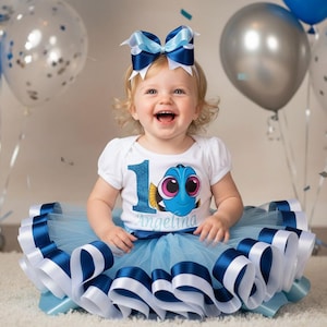 May include: A smiling toddler wearing a white bodysuit with a Dory graphic and the number 1. She is wearing a light blue tutu with white and navy blue ribbon trim and a matching bow. Balloons in blue and silver are in the background.