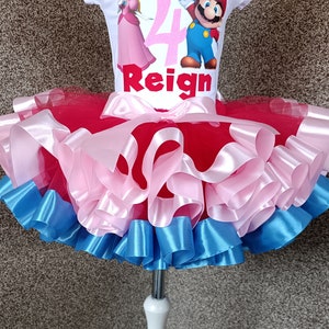 May include: A white t-shirt with a Super Mario theme, featuring Princess Peach and Mario, the number 4, and the name "Reign". The shirt is paired with a layered tutu skirt in red, pink, and blue.