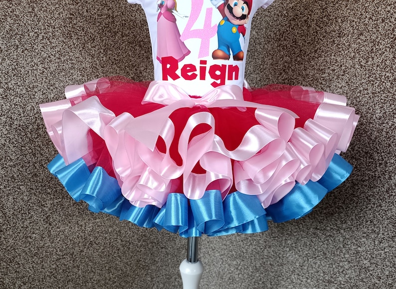 May include: A white t-shirt with a Super Mario theme, featuring Princess Peach and Mario, the number 4, and the name "Reign". The shirt is paired with a layered tutu skirt in red, pink, and blue.
