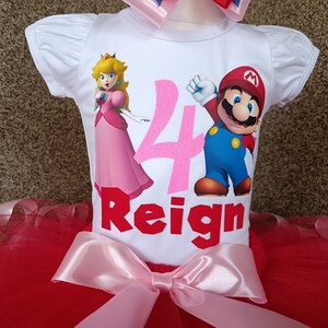May include: A white short-sleeved top featuring Princess Peach and Mario illustrations, the number 4, and the name "Reign" in red. It's combined with a red tutu skirt and a pink bow. A matching bow is at the top.