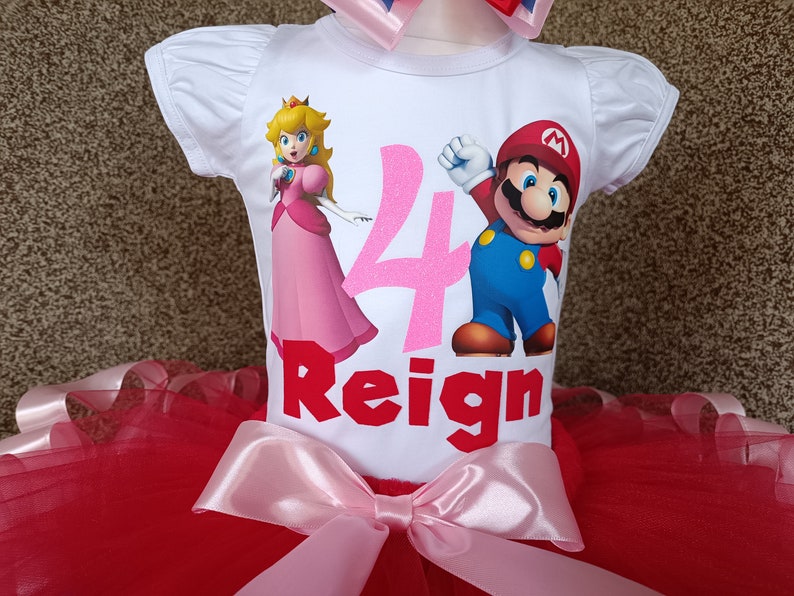 May include: A white short-sleeved top featuring Princess Peach and Mario illustrations, the number 4, and the name "Reign" in red. It's combined with a red tutu skirt and a pink bow. A matching bow is at the top.
