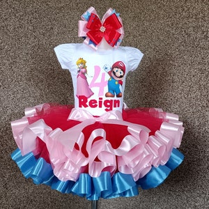 May include: A white t-shirt featuring Princess Peach and Mario, the number 4, and the name "Reign". The outfit includes a red, pink, and blue tutu skirt and a matching bow. A children's birthday outfit.