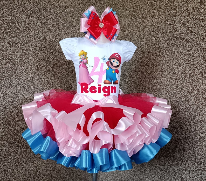 May include: A white t-shirt featuring Princess Peach and Mario, the number 4, and the name "Reign". The outfit includes a red, pink, and blue tutu skirt and a matching bow. A children's birthday outfit.