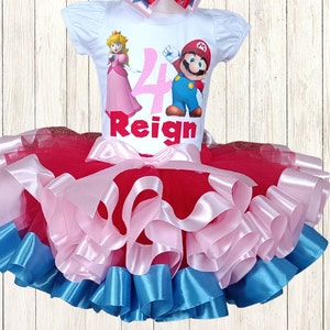 May include: A white t-shirt featuring Princess Peach and Mario, the number 4, and the name "Reign". The outfit includes a layered tutu skirt in red, pink, and blue. A matching bow is also included. The outfit is for a child.