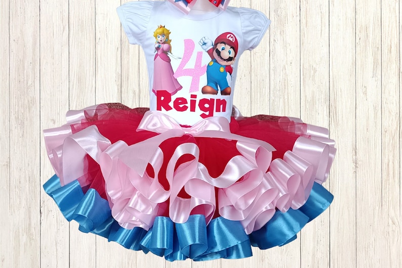 May include: A white t-shirt featuring Princess Peach and Mario, the number 4, and the name "Reign". The outfit includes a layered tutu skirt in red, pink, and blue. A matching bow is also included. The outfit is for a child.