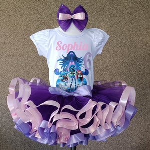 May include: A white short-sleeved shirt with the name "Sophia" and the number "6" printed in pink. The shirt features a colorful graphic of animated characters. A purple tutu skirt with layers of purple, lavender, and pink ribbons and a matching bow complete the outfit.