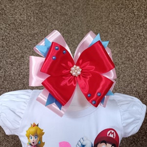 May include: A large, layered hair bow in red, pink, and blue. The bow features a central embellishment of pearls and blue rhinestones. The bow is attached to a white shirt with cartoon characters.