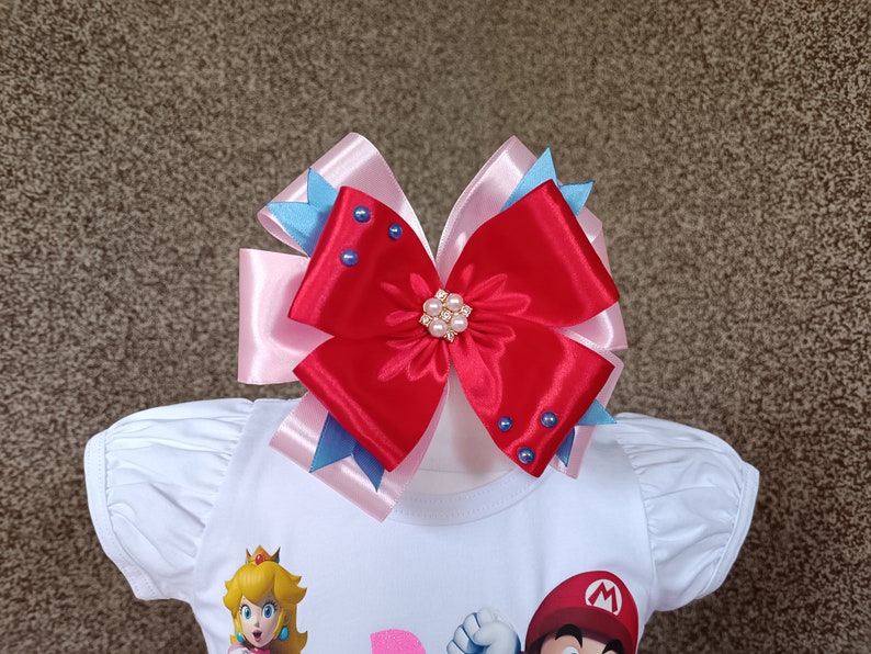 May include: A large, layered hair bow in red, pink, and blue. The bow features a central embellishment of pearls and blue rhinestones. The bow is attached to a white shirt with cartoon characters.