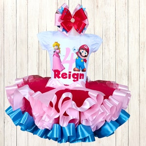 May include: A white short-sleeved top featuring Princess Peach and Mario, the number 4, and the name "Reign". The top is combined with a layered tutu skirt in red, pink, and blue. A large bow with red, pink, and blue ribbons completes the outfit.