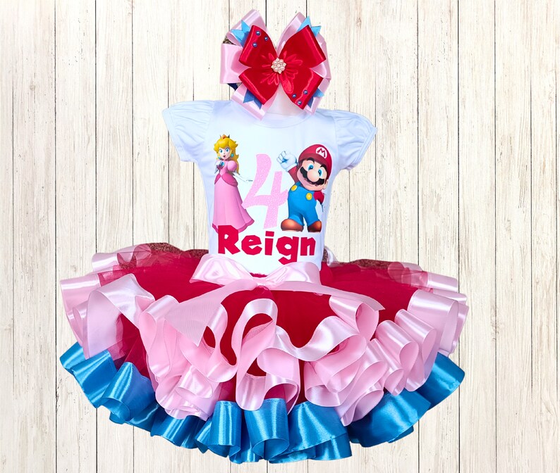May include: A white short-sleeved top featuring Princess Peach and Mario, the number 4, and the name "Reign". The top is combined with a layered tutu skirt in red, pink, and blue. A large bow with red, pink, and blue ribbons completes the outfit.