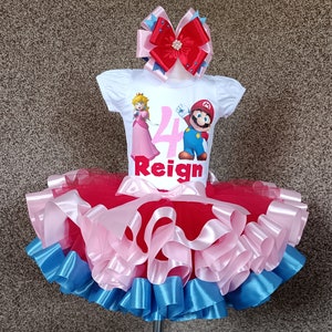 May include: A white t-shirt featuring Princess Peach and Mario, with the number 4 and the name "Reign". The outfit includes a red, pink, and blue tutu and a matching bow, suitable for a child's birthday.