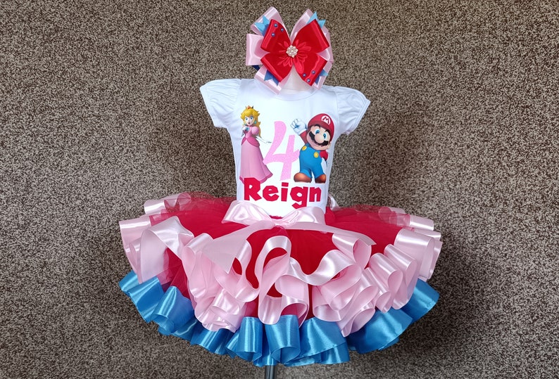 May include: A white t-shirt featuring Princess Peach and Mario, with the number 4 and the name "Reign". The outfit includes a red, pink, and blue tutu and a matching bow, suitable for a child's birthday.