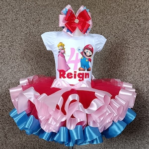 May include: A white t-shirt featuring Princess Peach and Mario, the number 4, and the name "Reign". The outfit includes a red, pink, and blue tutu skirt and a matching bow, perfect for a birthday.