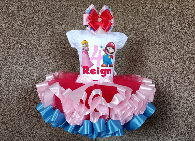 May include: A white t-shirt featuring Princess Peach and Mario, the number 4, and the name "Reign". The outfit includes a red, pink, and blue tutu skirt and a matching bow, perfect for a birthday.