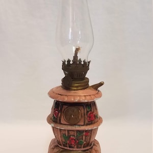May include: A pink ceramic oil lamp shaped like a wood-burning stove with a glass chimney and floral details. The stove has a decorative metal door and a base with four legs.