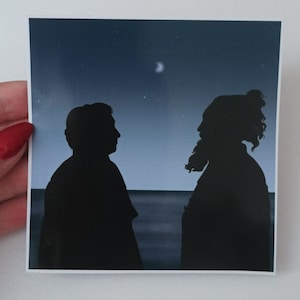 May include: Silhouette of two people standing side by side against a dark blue sky with a crescent moon and stars. The figures are facing away from each other, with the person on the left looking to the right and the person on the right looking to the left.