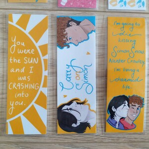 May include: Three yellow bookmarks with different designs. The first bookmark has a yellow sun with the text "You were the SUN and I was CRASHING into you." The second bookmark has a blue and white design with the text "Carry on Simon." The third bookmark has a yellow background with the text "I'm going to die kissing Simon Snow. Aleister Crowley. I'm living a charmed life."