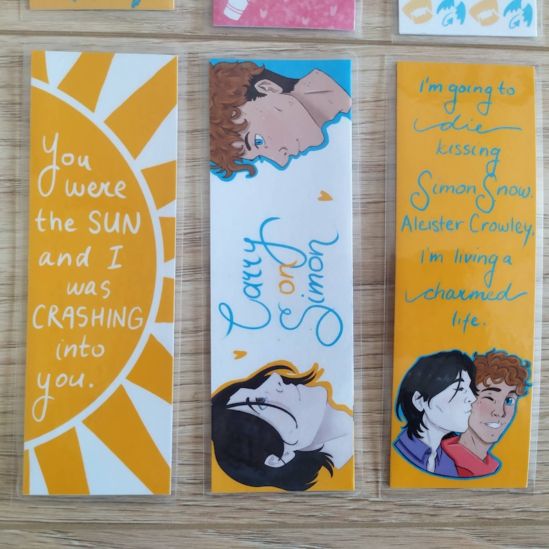 May include: Three yellow bookmarks with different designs. The first bookmark has a yellow sun with the text "You were the SUN and I was CRASHING into you." The second bookmark has a blue and white design with the text "Carry on Simon." The third bookmark has a yellow background with the text "I'm going to die kissing Simon Snow. Aleister Crowley. I'm living a charmed life."