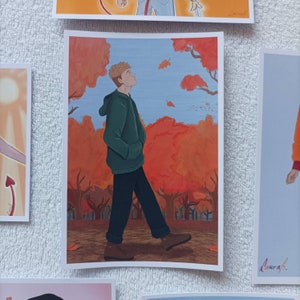 Puede incluir: Un hombre con una sudadera con capucha verde y pantalones vaqueros azules camina por un parque con follaje oto&ntilde;al rojo y naranja. El cielo es azul y hay hojas cayendo de los &aacute;rboles.
