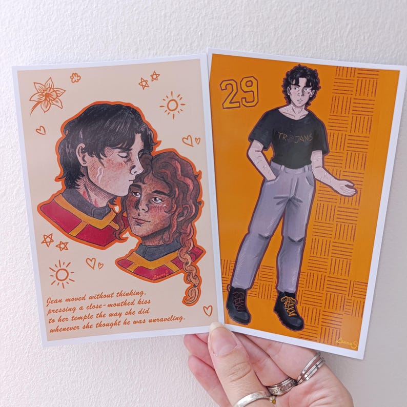 May include: Two art prints. One shows a couple kissing with text, the other a person with the number 29. The first print has a beige background with a couple kissing, with text below. The second print has an orange background with a person wearing a black shirt.