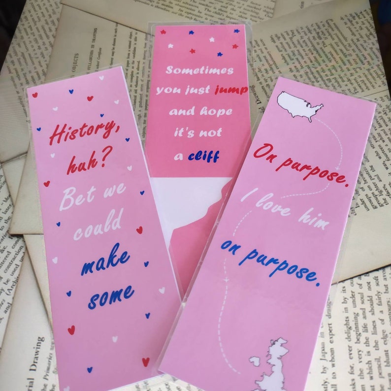 May include: Three pink bookmarks with different quotes and designs. The first bookmark says "History, huh? Bet we could make some." The second bookmark says "Sometimes you just jump and hope it's not a cliff." The third bookmark says "On purpose. I love him on purpose."