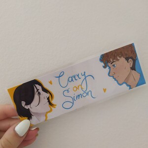May include: A white bookmark with the text "Carry on Simon" in blue cursive writing. The bookmark features illustrations of two people, one with brown hair and the other with blonde hair, looking at each other.
